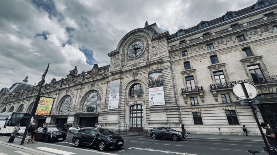 Exterior view of the Musée d'Orsay taken from the River Seine. With sign and large black clock. Several black taxis are parked in the foreground. 2